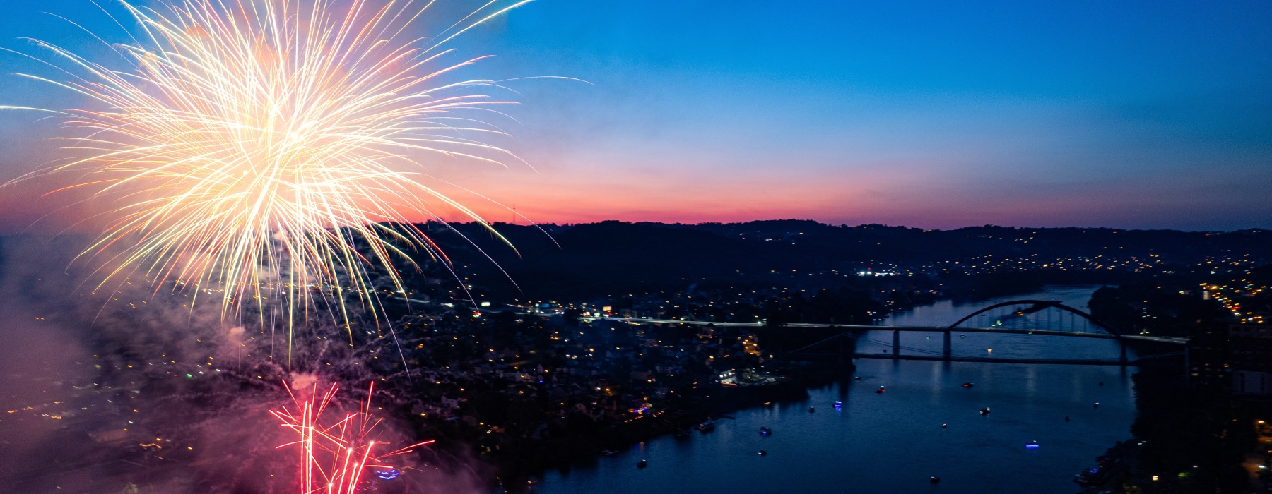 Fourth of July fireworks lighting up the sky at sunset over a river and bridge