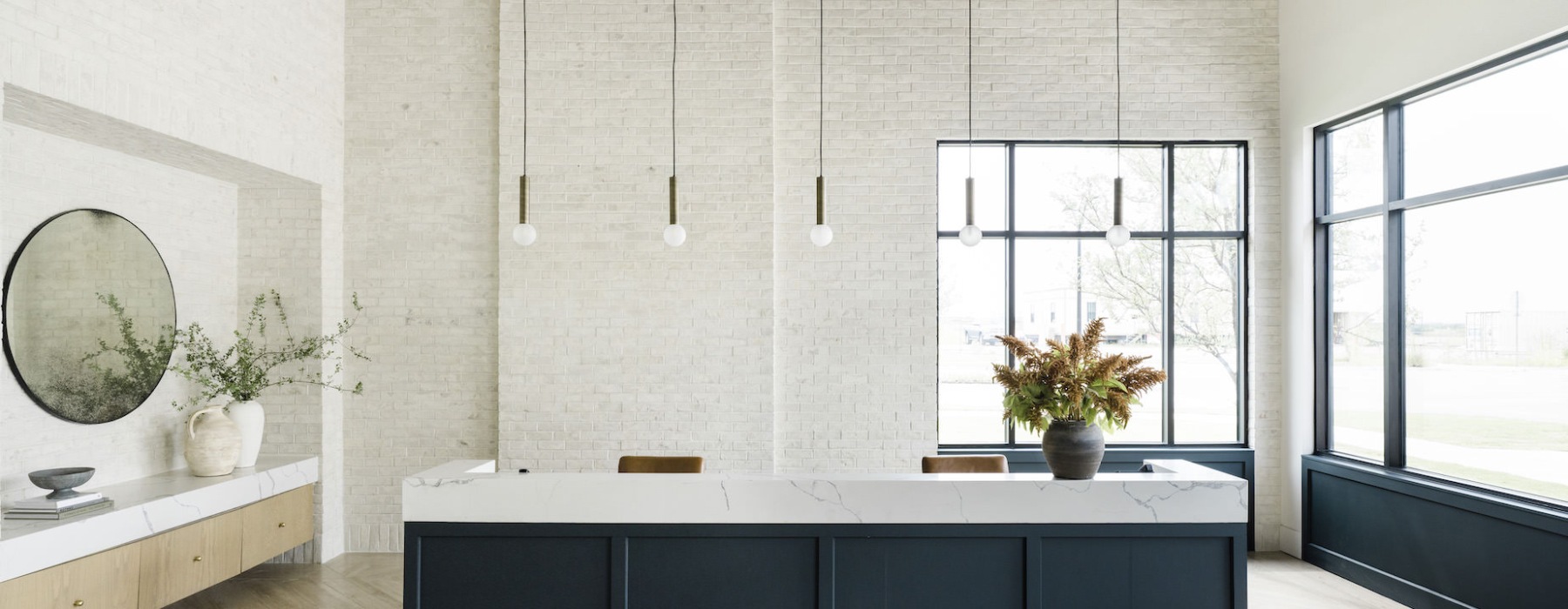 A modern, minimalist reception area with a long, marble-topped counter and black base. The counter has a large vase with dried orange flowers. Behind it, there is a circular mirror and potted plants on a side table. Pendant lights hang from the ceiling.