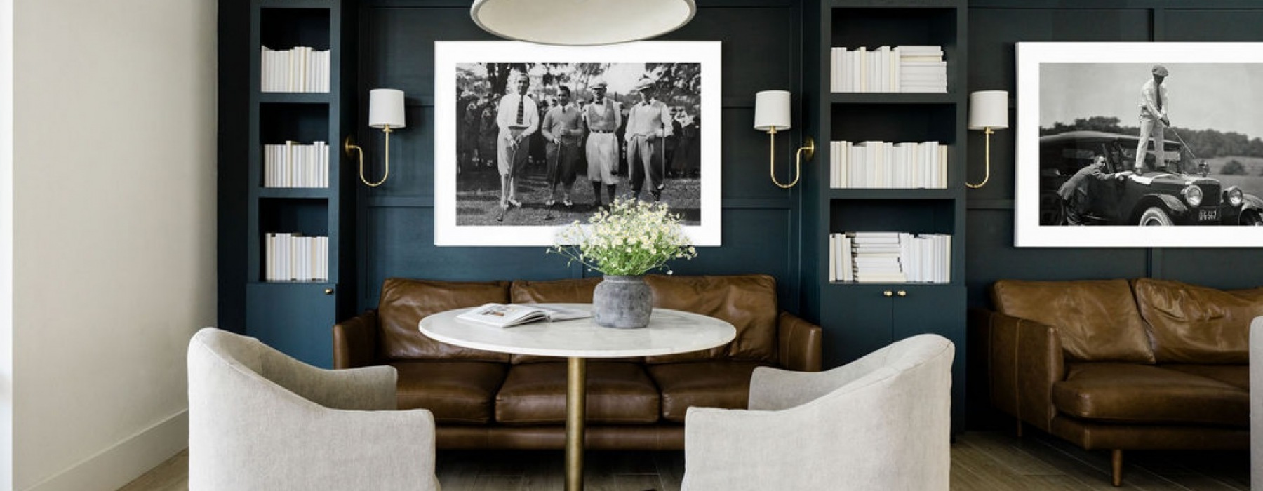 Comfortable lounge area with dark blue bookshelves, brown leather couches, a white marble table, and beige chairs. Framed black-and-white photos of historical scenes on the wall.