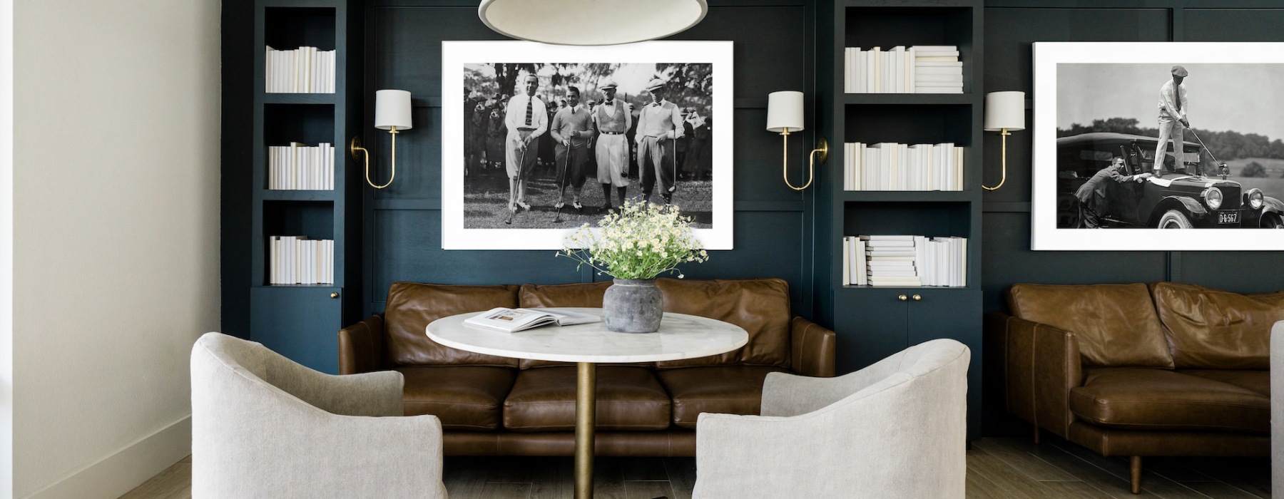 Chic lounge area with leather sofas, a round marble table, and upholstered chairs, set against a dark built-in bookshelf with white books and black-and-white vintage photos, creating a modern yet classic atmosphere.