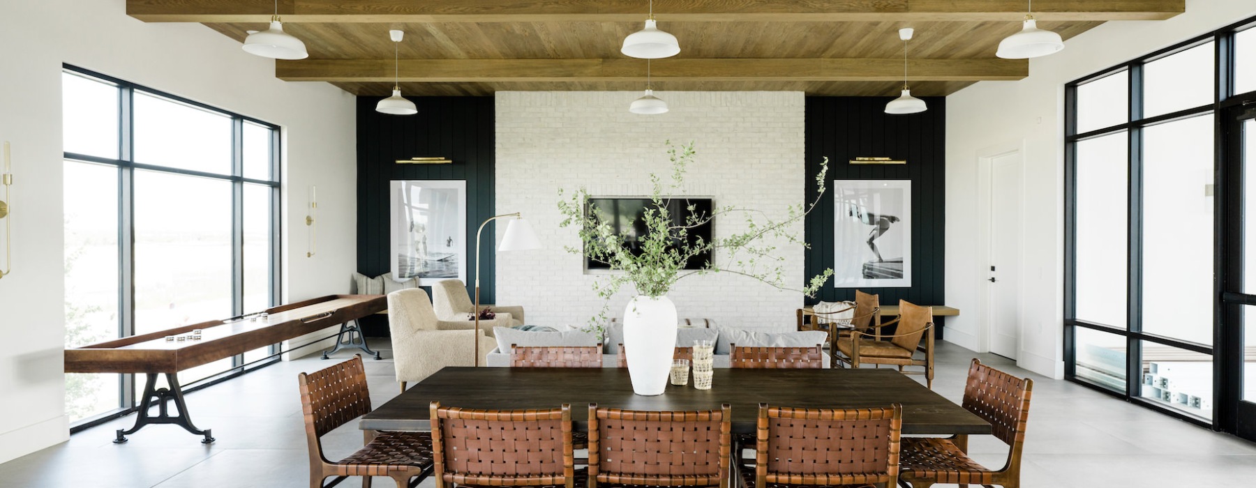 A modern, airy living and dining room with large windows. The dining area has a dark wooden table and woven leather chairs. The living area features a white sofa, armchairs, and a TV on a light brick wall. A shuffleboard table is along the left wall.