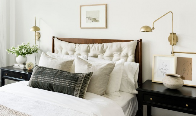 Stylish bedroom with a wooden headboard and tufted pillows, featuring neutral-toned bedding, black nightstands with brass accents, and minimalist wall art in a bright, modern space.