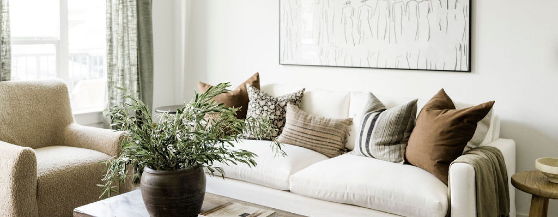 Bright and cozy living room with a white couch adorned with earth-tone pillows, a potted plant on a wooden coffee table, and a soft beige armchair next to large windows with natural light.