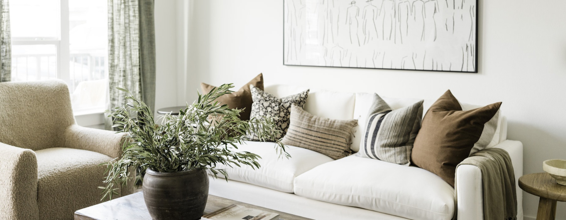 Bright and cozy living room with a white couch adorned with earth-tone pillows, a potted plant on a wooden coffee table, and a soft beige armchair next to large windows with natural light.