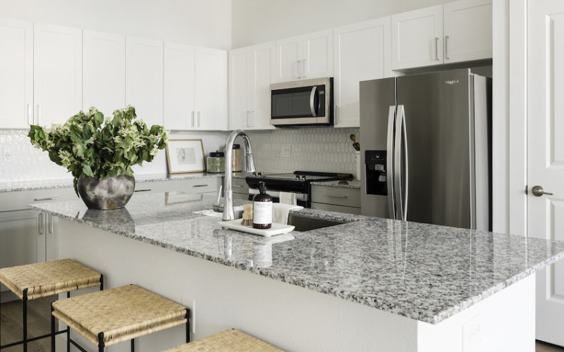 Modern kitchen with a large granite island, three wicker stools, stainless steel appliances, white cabinetry, a vase with green plants, and a framed picture on the counter.