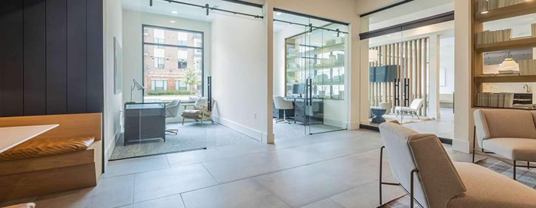 A modern and open work area featuring glass-walled offices and seating areas. The space has a bright, clean design with natural light coming through large windows, and minimalistic furniture such as armchairs and a small desk setup. Warm wood tones and so
