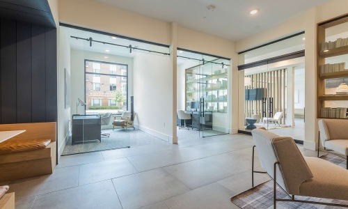 A modern and open work area featuring glass-walled offices and seating areas. The space has a bright, clean design with natural light coming through large windows, and minimalistic furniture such as armchairs and a small desk setup. Warm wood tones and so