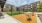 A modern apartment complex with multiple levels and balconies surrounds a central courtyard featuring a well-manicured green lawn. The courtyard is lined with gravel paths, outdoor seating, and lamp posts, creating a communal space. The sky is clear with 