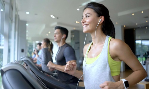 A woman with a ponytail and earphones runs on a treadmill in a gym, smiling and looking ahead. She is wearing a gray tank top over a yellow sports bra. In the background, other people are also using treadmills, and large windows let in natural light.