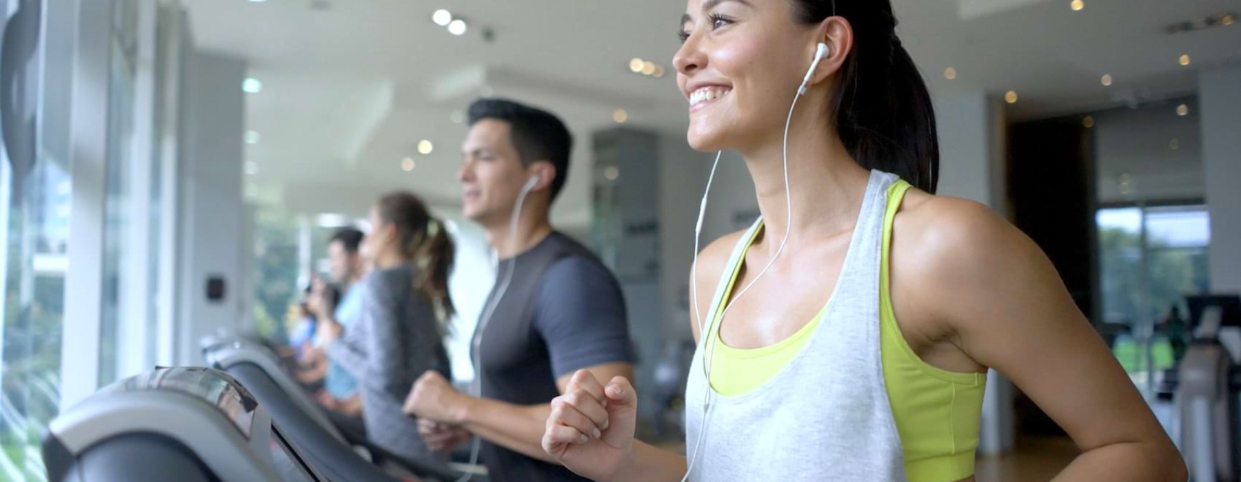 A woman with a ponytail and earphones runs on a treadmill in a gym, smiling and looking ahead. She is wearing a gray tank top over a yellow sports bra. In the background, other people are also using treadmills, and large windows let in natural light.