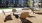 A modern outdoor courtyard features four wicker chairs around a round table on a gravel surface. Nearby, two hammocks are set up on stands. In the background, greenery and a multi-story apartment building with balconies are visible.